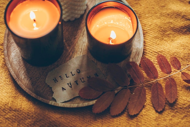 Bandeja de madera con velas encendidas y tarjeta con texto hola otoño sobre cuadros de lana naranja en la cama Otoño