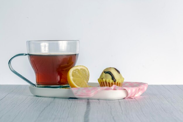 Foto bandeja en una mesa de madera que contiene una taza de té acompañada de una rodaja de limón y un pastelito pequeño