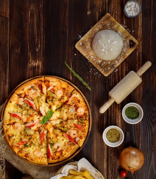 Bandeja de pizza de mariscos casera rodeada de ingredientes ubicada en fila debajo del fondo de madera y decorada con plantas secas, bebida fría y papas fritas con espacio de copia.