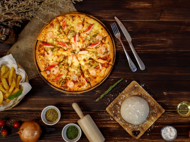 Bandeja de pizza de mariscos casera rodeada de ingredientes ubicada en fila debajo del fondo de madera y decorada con plantas secas, bebida fría y papas fritas con espacio de copia.