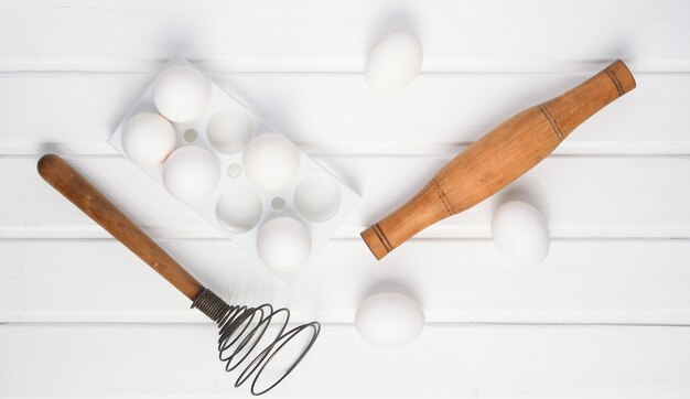Bandeja plástica com ovos brancos, rolo e bata em uma mesa de madeira branca
