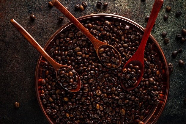 Bandeja redonda de cobre, cucharas de madera llenas de granos de café kopi luwak en superficie oscura, vista superior