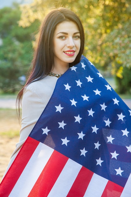 Bandera americana y mujer (4 de julio). Hermosa mujer joven con vestido clásico sosteniendo la bandera americana en el parque. modelo de moda abrazándonos sonriendo y mirando a la cámara. estilo de vida de EE. UU ..