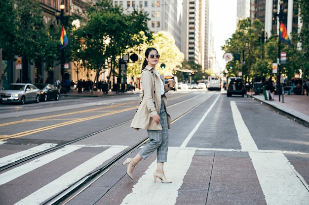 bandera del arco iris colgada en la calle de fondo en la ciudad de san francisco urbano. joven universitaria sosteniendo una taza de café yendo a la universidad caminando por el paso de cebra. mujer asiática con gafas de sol en verano.