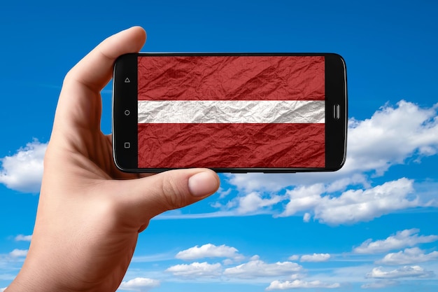 Bandera de Letonia en la pantalla del teléfono El teléfono inteligente en la mano muestra una bandera sobre un fondo de cielo con nubes Fotografía móvil