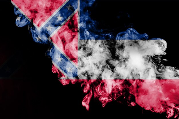 Foto la bandera nacional del estado estadounidense de mississippi contra un humo gris en el día de la independencia en diferentes colores azul rojo y amarillo disputas políticas y religiosas costumbres y entrega