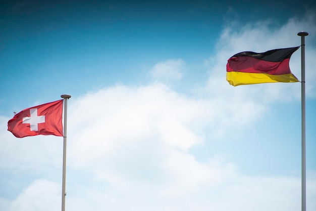 Banderas ondeantes de Suiza y Alemania en el poste de metal contra el cielo azul nublado