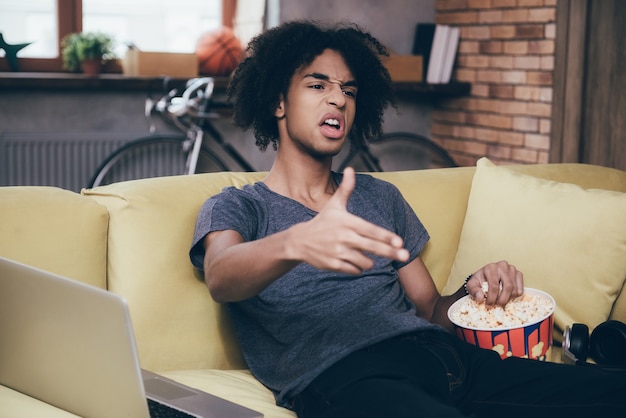 Bang! Jovem africano assistindo TV e gesticulando com o dedo enquanto está sentado com um balde de pipoca no sofá de casa