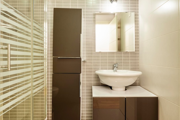 Banheiro com pequena cômoda branca com portas marrons com espelho quadrado sem moldura e pia de porcelana de coluna combinando e cabine de chuveiro com portas de vidro deslizantes