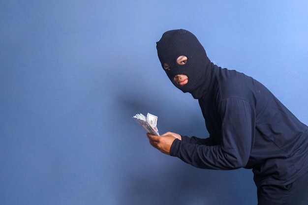 Foto bankräuber in einer balaclava hält einen haufen gestohlener dollars auf blauem studiohintergrund isoliert