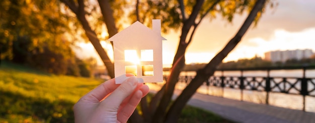 Banner con una casa de papel en una mano contra el cielo del atardecer. concepto de hipoteca, agente inmobiliario, hipoteca