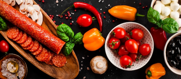 Banner da Web com ingredientes de pizza no fundo cinza escuro. Calabresa, queijo mussarela, tomate, azeitonas, cogumelos e farinha são produtos diferenciados para a confecção de pizzas e massas