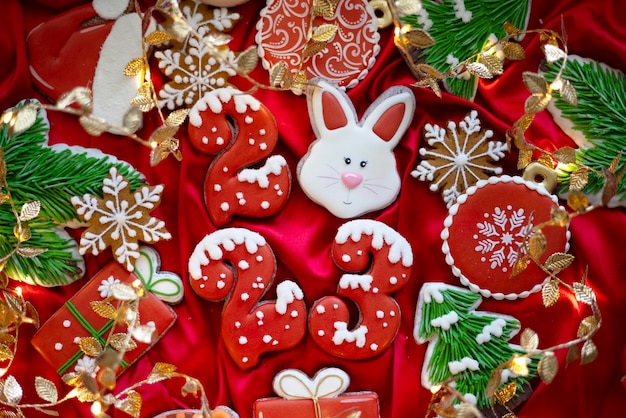 Banner für Weihnachten und Neujahr Lebkuchen Zahlen 2023 Schneeflocken Weihnachtsmütze Weihnachtsbäume Girlanden auf rotem Seidenstoff Hintergrund Lebkuchen-Kaninchen-Symbol Chinesischer Tierkreiskalender