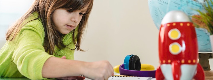 Foto banner horizontal o encabezado con una niña que se divierte dibujando con colores de cera en un cuaderno escolar para la tarea en el escritorio hay un globo y un cohete rojo para viajes espaciales