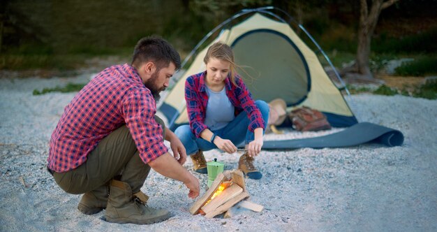 Banner-Mann beim Camping, der Brennholz auf dem Land anzündet, mit Frau in der Nähe des Zeltes