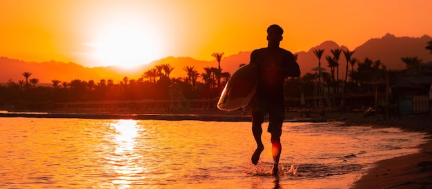 Banner Silhouette Mann Surfer im Abendlicht läuft Strand hinunter mit Surfbrett in der Nähe von Gewässern und genießt Sommerzeit-Praxis-Hobby während der Kurzurlaube Typ, der eine tropische Insel mit Wellen nachbildet