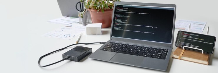 Foto banner del sitio web de computadora portátil de grafito y teléfono inteligente negro con códigos en una mesa de trabajo de madera blanca