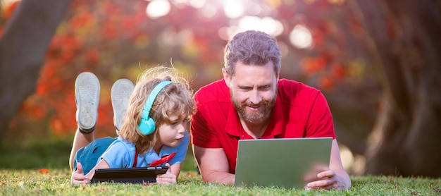 Banner von Vater auf Sohn Schuljunge mit Laptop studieren online auf Gras liegend Kindheit und Elternschaft glücklicher Junge hört Musik in Kopfhörern Webinar-Videolektion