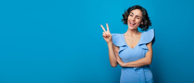 Bannerfoto einer aufgeregten, glücklichen, süßen Frau in einem langen blauen Kleid, die in die Kamera schaut, während sie mit einem strahlenden Lächeln posiert und ein Siegeszeichen auf der Kamera zeigt