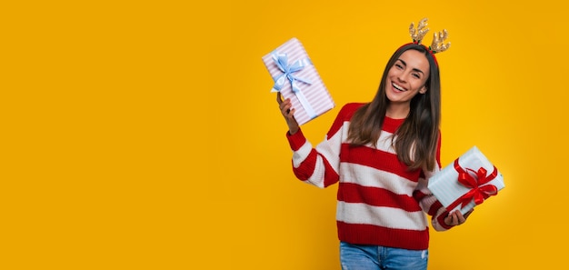 Bannerfoto einer glücklichen aufgeregten jungen brünetten Frau mit Hirschhut, mit vielen bunten Weihnachtsgeschenkboxen in den Händen einzeln auf gelbem Hintergrund.