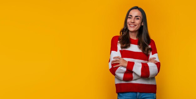 Bannerfoto einer süßen lächelnden jungen Frau in einem Pullover mit gekreuzten Händen, während sie isoliert auf gelbem Hintergrund posiert