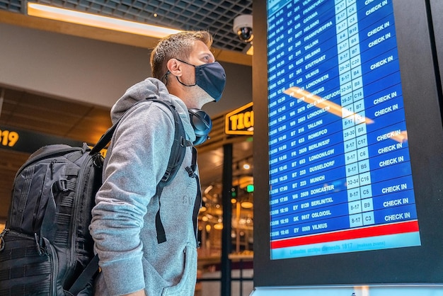 Bannerfoto eines jungen Mannes mit Gesichtsmaske mit Koffer, der den Flugannullierungsstatus auf der Flughafeninformationstafel im leeren Flughafen überprüft. Airline-Pleite, Airline-Krise oder neues Normalkonzept