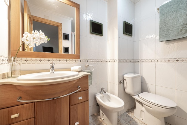 Baño con armarios de madera con puertas y cajoneras un espejo enmarcado en la pared una flor un toallero integrado al mueble y sanitarios blancos