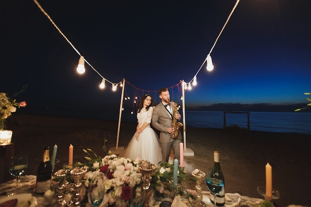 Banquete de bodas en la orilla del mar por la noche. El novio toca el saxofón.