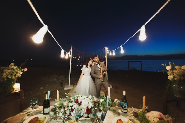 Banquete de bodas en la orilla del mar por la noche. El novio toca el saxofón.