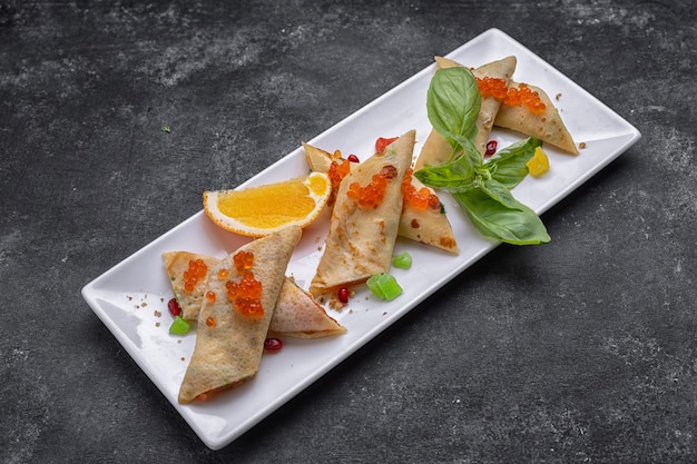 Banquete de panqueques con caviar rojo y salmón, sobre un plato rectangular blanco, con hojas de albahaca y una rodaja de naranja