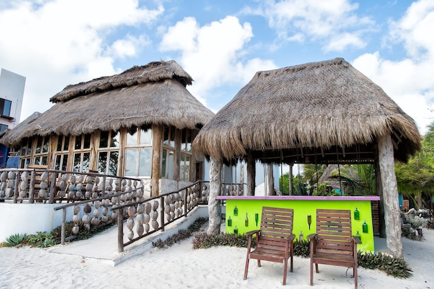 Bar de praia exótico com construção de casas e telhado de palha perto de cadeiras na areia ao ar livre no fundo do céu azul nublado