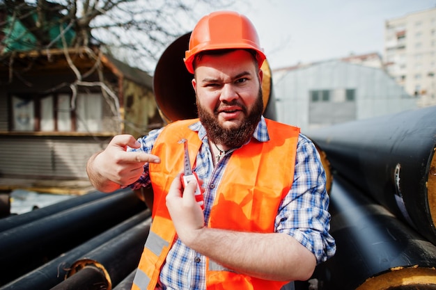 Barba divertida trabajador hombre traje trabajador de la construcción en casco naranja de seguridad cerca de tubos de acero con cinta métrica a mano muestra pequeño tamaño Concepto de pene pequeño