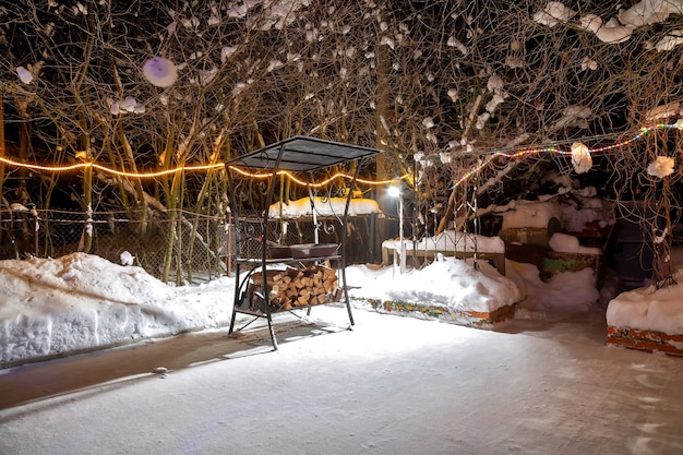 Barbacoa cerca de la casa en invierno. De noche, las guirnaldas arden, está nevando. Patio de juegos vacío. Colocan leña cosechada debajo de la parrilla.