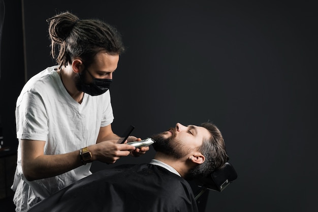 Barbear a barba na barbearia. Barbeiro com dreadlocks em máscara médica preta aparar homem barbudo em quarentena coronavírus covid-19.
