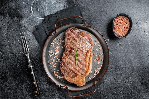 Barbecue Gegrilltes Roastbeef oder New Yorker Rindersteak mit rosa Salz und Kräutern Schwarzer Hintergrund Draufsicht