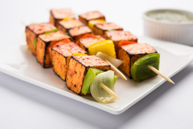 Barbecue Paneer Tikka Kabab - cubos de requesón indio Tandoori en salsa roja y blanca en palitos, servidos en un plato con chutney verde sobre una mesa colorida o de madera, enfoque selectivo