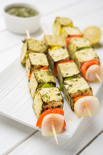 Barbecue Paneer Tikka Kabab - cubos de requesón indio Tandoori en salsa roja y blanca en palitos, servidos en un plato con chutney verde sobre una mesa colorida o de madera, enfoque selectivo