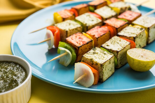 Barbecue Paneer Tikka Kabab - cubos de requesón indio Tandoori en salsa roja y blanca en palitos, servidos en un plato con chutney verde sobre una mesa colorida o de madera, enfoque selectivo