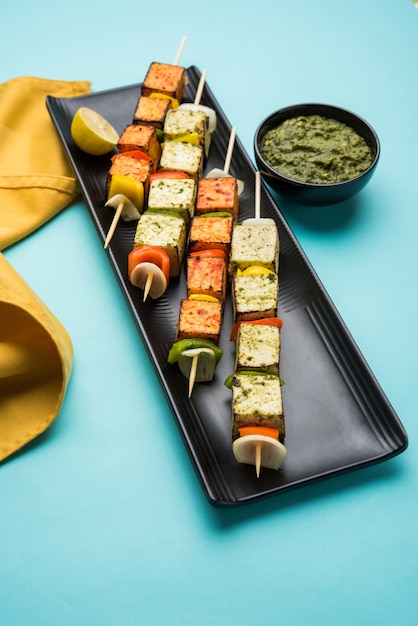 Barbecue Paneer Tikka Kabab - cubos de requesón indio Tandoori en salsa roja y blanca en palitos, servidos en un plato con chutney verde sobre una mesa colorida o de madera, enfoque selectivo