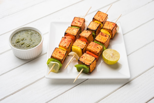 Barbecue Paneer Tikka Kabab - Indische Tandoori Hüttenkäsewürfel in roter und weißer Sauce in Stäbchen, serviert in einem Teller mit grünem Chutney über bunter oder hölzerner Tischplatte, selektiver Fokus