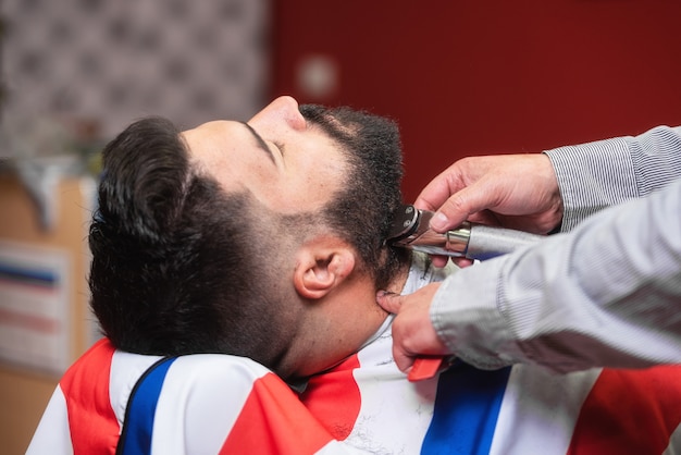 Barbeiro barbeando a barba de um homem barbudo bonito com um barbeador elétrico na barbearia.