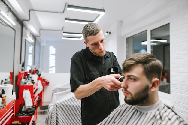 Barbeiro concentrado faz cliente barbudo de cabelo em uma barbearia moderna e leve usa um aparador