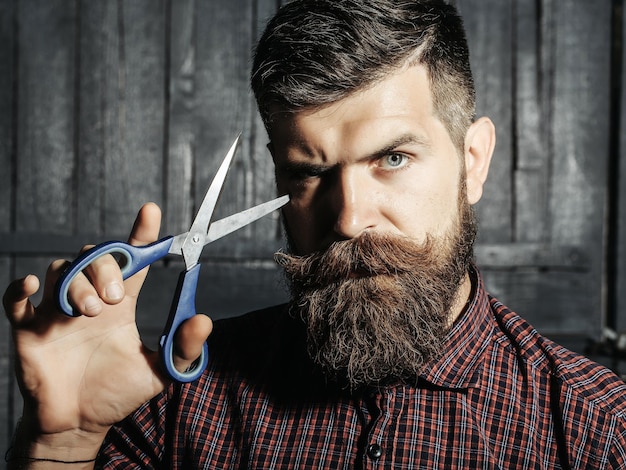 Barbeiro hipster homem barbudo com barba e bigode na camisa segura tesoura afiada e cabelo cortado no salão de cabeleireiro em fundo de madeira