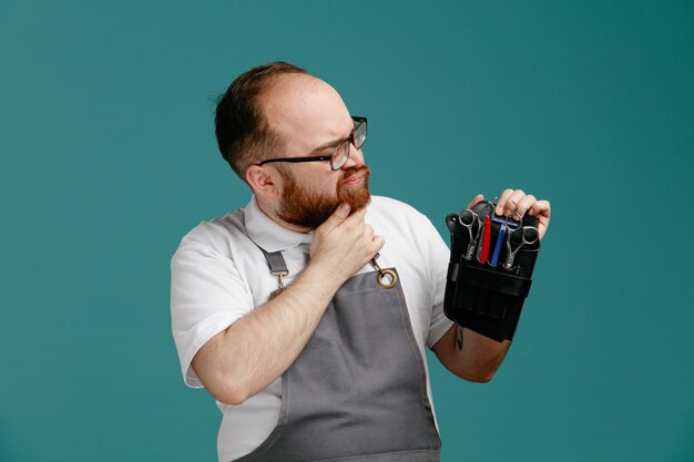 Barbeiro jovem inseguro vestindo uniforme e óculos segurando e olhando para o saco de barbeiro com ferramentas de barbeiro, mantendo a mão no queixo isolado no fundo azul