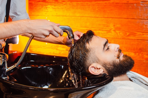 Barbeiro lava a cabeça de um jovem bonitão com barba e bigode na pia