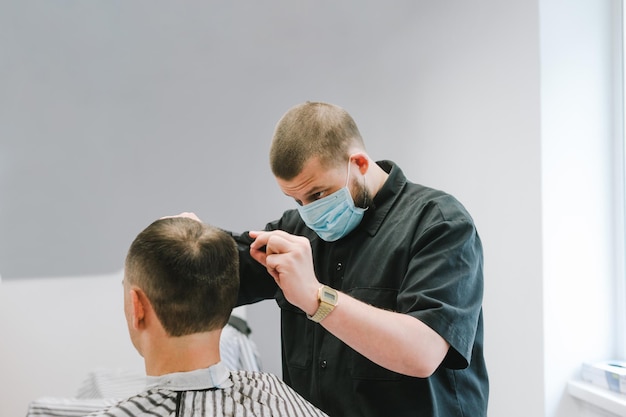 Barbeiro profissional em uma máscara médica corta o cabelo do cliente em uma barbearia masculina Trabalha em cabeleireiros em quarentena COVID19