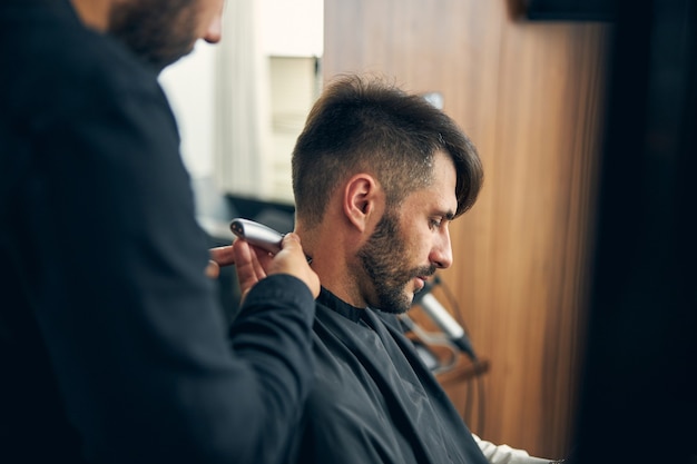 Barbeiro profissional usando o cortador de cabelo enquanto cria um corte de cabelo elegante, ficando em primeiro plano