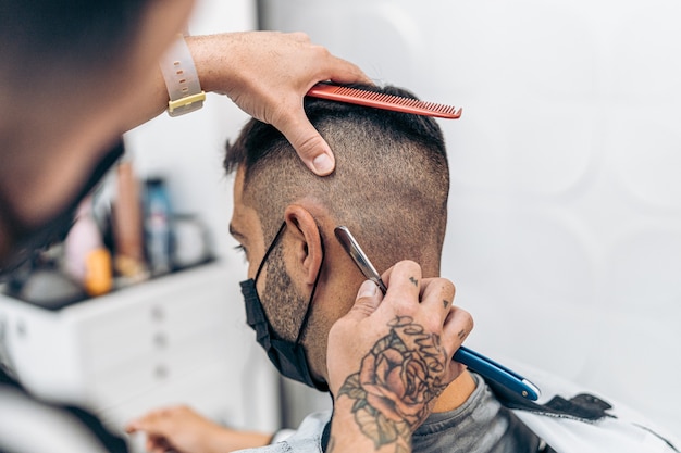 Barbeiro raspando o cabelo de um homem caucasiano sentado em uma cadeira usando uma navalha em uma barbearia