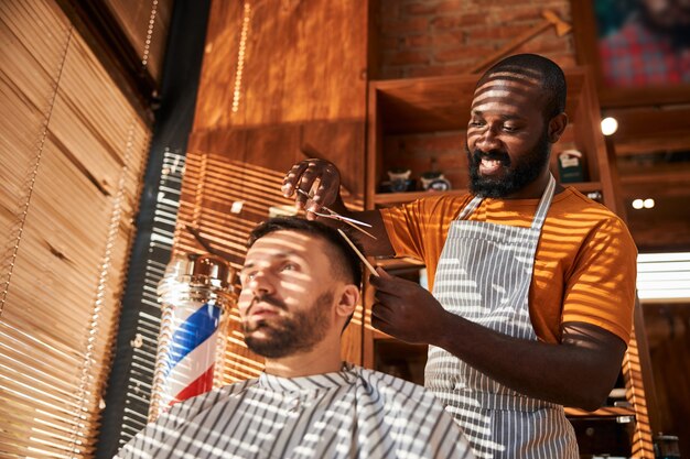 Barbeiro sorridente cortando o cabelo do cliente com uma tesoura na barbearia