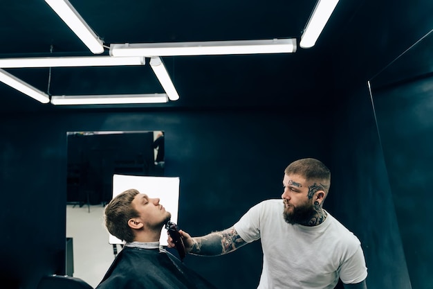Barbeiro tatuado aparando homem barbudo com máquina de barbear na barbearia Processo de penteado Cabeleireiro cortando a barba de um homem barbudo